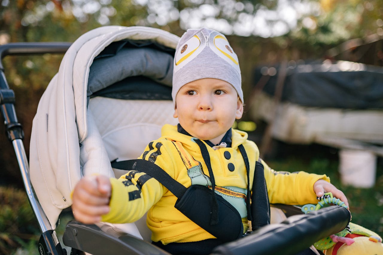 A cute baby sitting in a stroller outdoors, dressed in a yellow jacket and a bonnet.