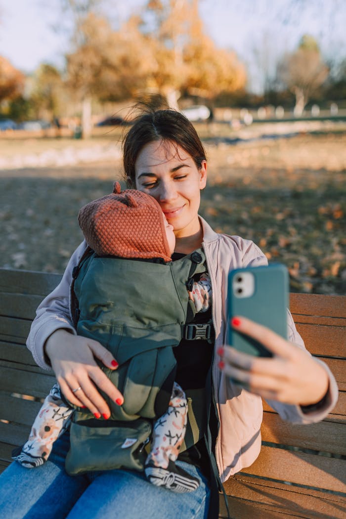 A young woman takes a selfie while holding her baby in a carrier at the park.