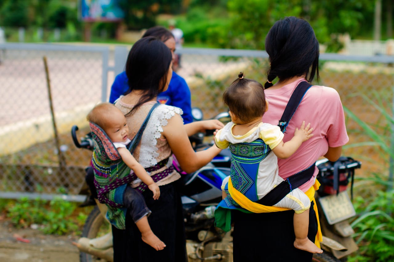 Two mothers with babies in traditional carriers riding on a scooter outdoors.
